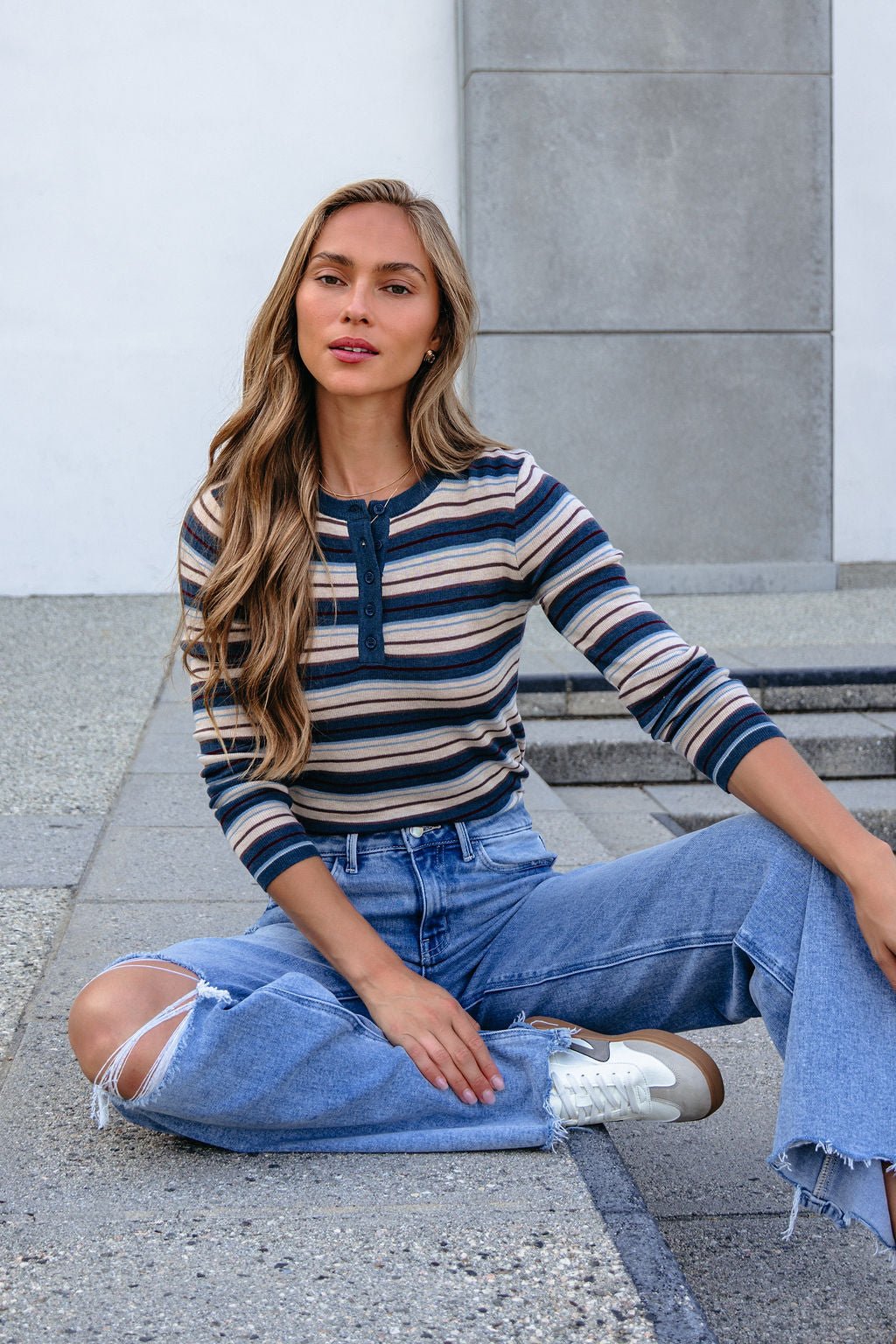 Woman with long hair wears a Teal Striped Henley Long Sleeve Top and sits cross-legged on concrete outdoors.