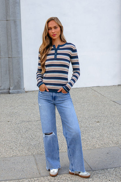 Woman in a Teal Striped Henley Long Sleeve Top and ripped jeans stands on pavement by a white wall and stone pillar.