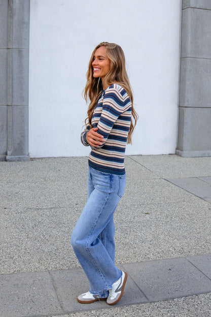Woman with long hair smiles, arms crossed, wearing a Teal Striped Henley Long Sleeve Top and jeans by a white and gray building.