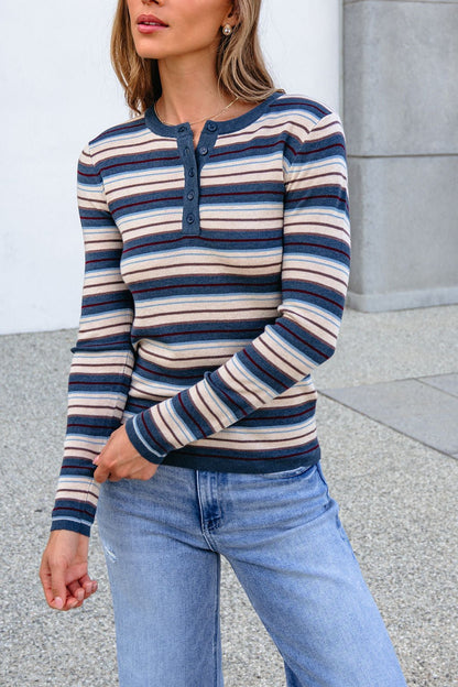 Woman in a Teal Striped Henley Long Sleeve Top and light blue jeans stands by a concrete building.