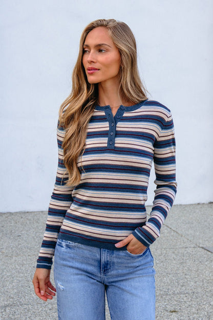 Woman with long hair wears the Teal Striped Henley Long Sleeve Top and jeans, standing outside against a light background.