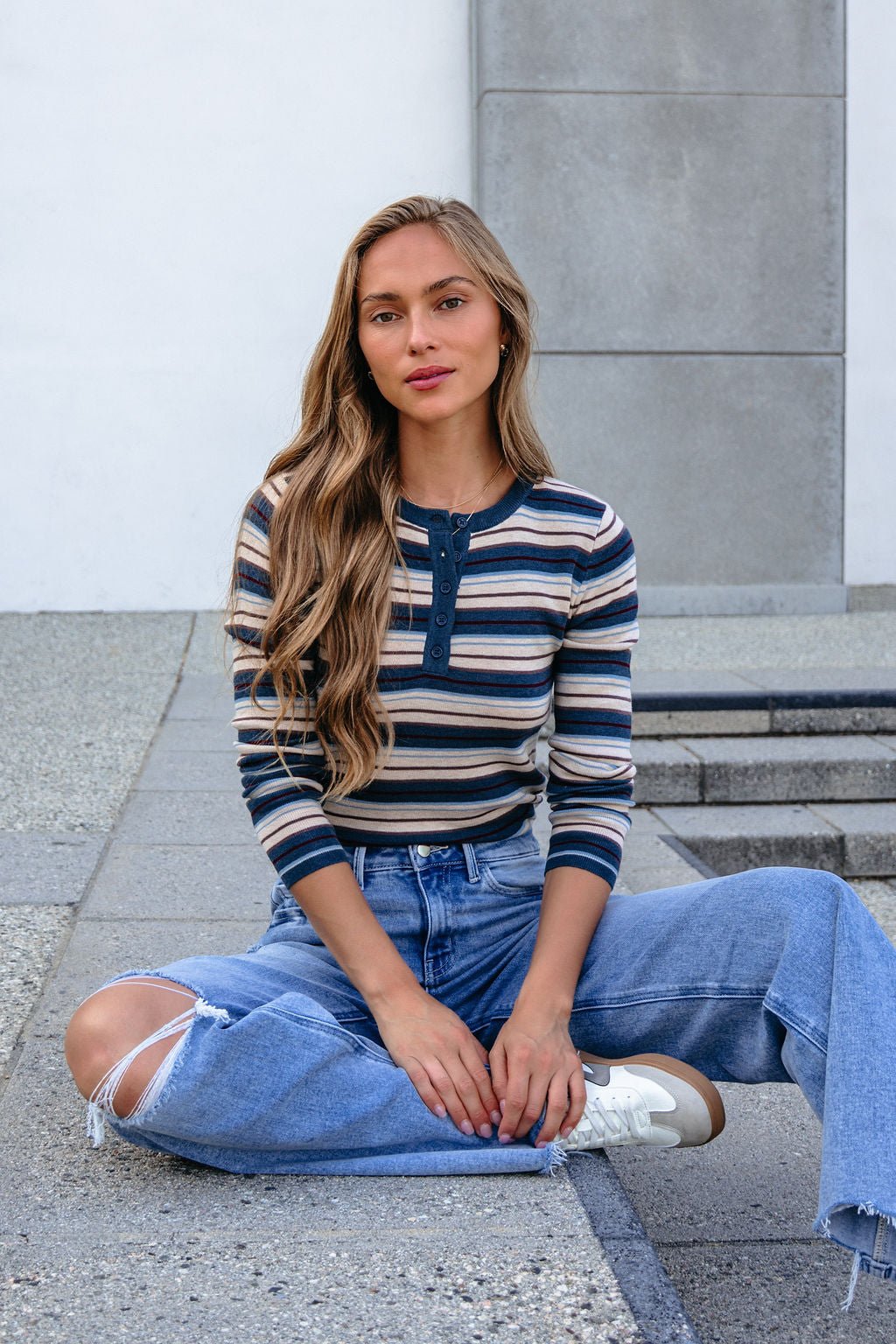 A woman wears the Teal Striped Henley Long Sleeve Top and ripped jeans, sitting cross-legged on stone, looking at the camera.