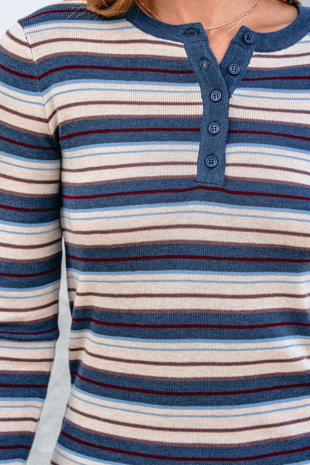 Someone wears the Teal Striped Henley Long Sleeve Top featuring a relaxed fit and four blue, cream, and maroon button front details.