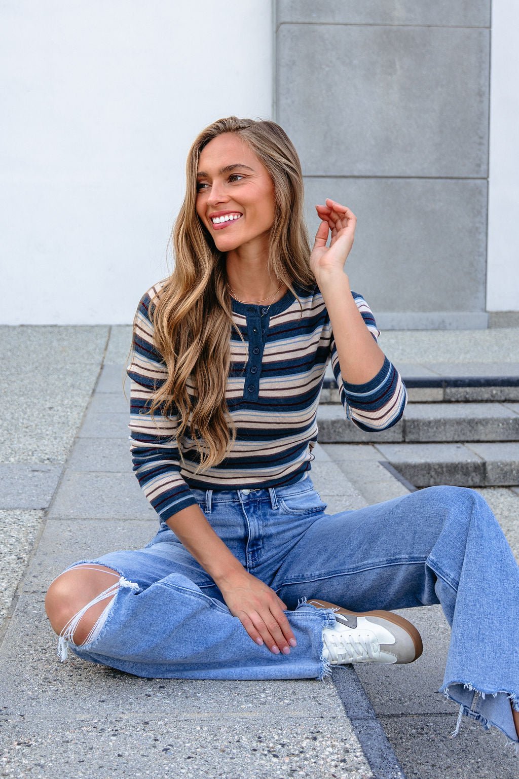 Woman with long wavy hair smiles, sitting cross-legged in ripped jeans and a Teal Striped Henley Long Sleeve Top on concrete steps.