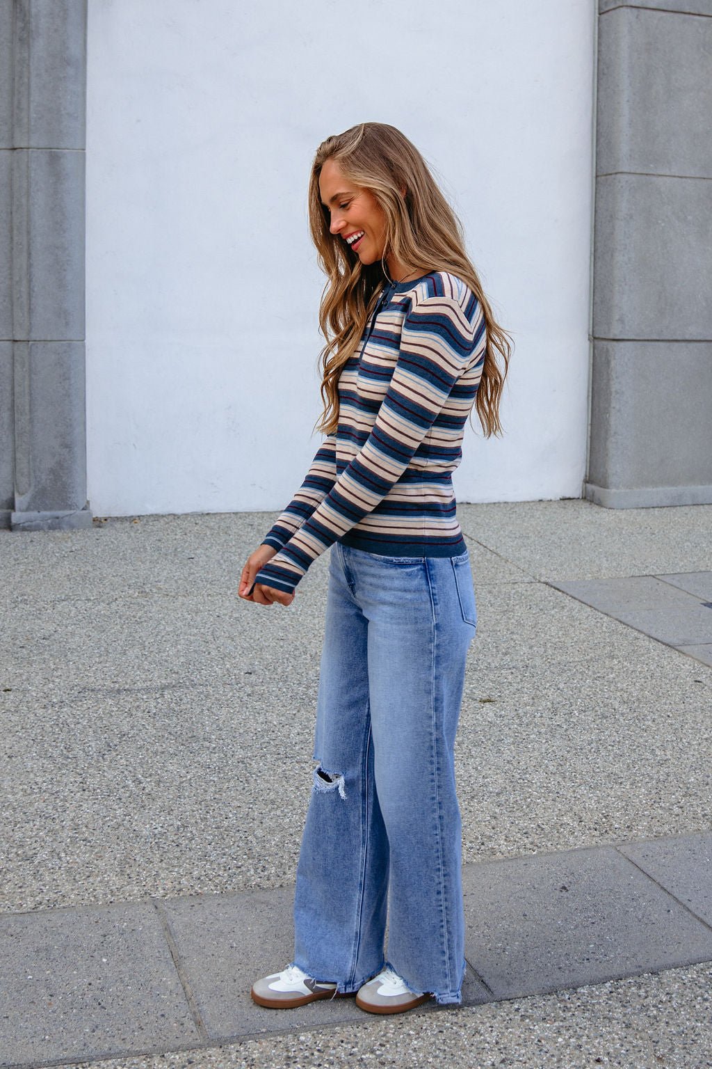 Smiling woman outdoors in a Teal Striped Henley Long Sleeve Top and wide-leg ripped jeans stands on a paved surface.
