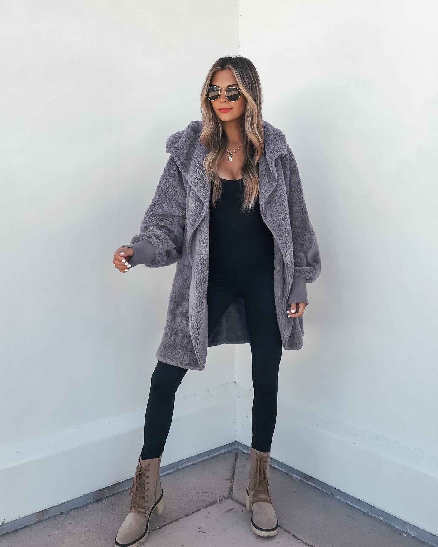 Woman in sunglasses, a gray Teddy Fleece Hooded Cardigan, black leggings, and tan lace-up boots stands by a white wall.
