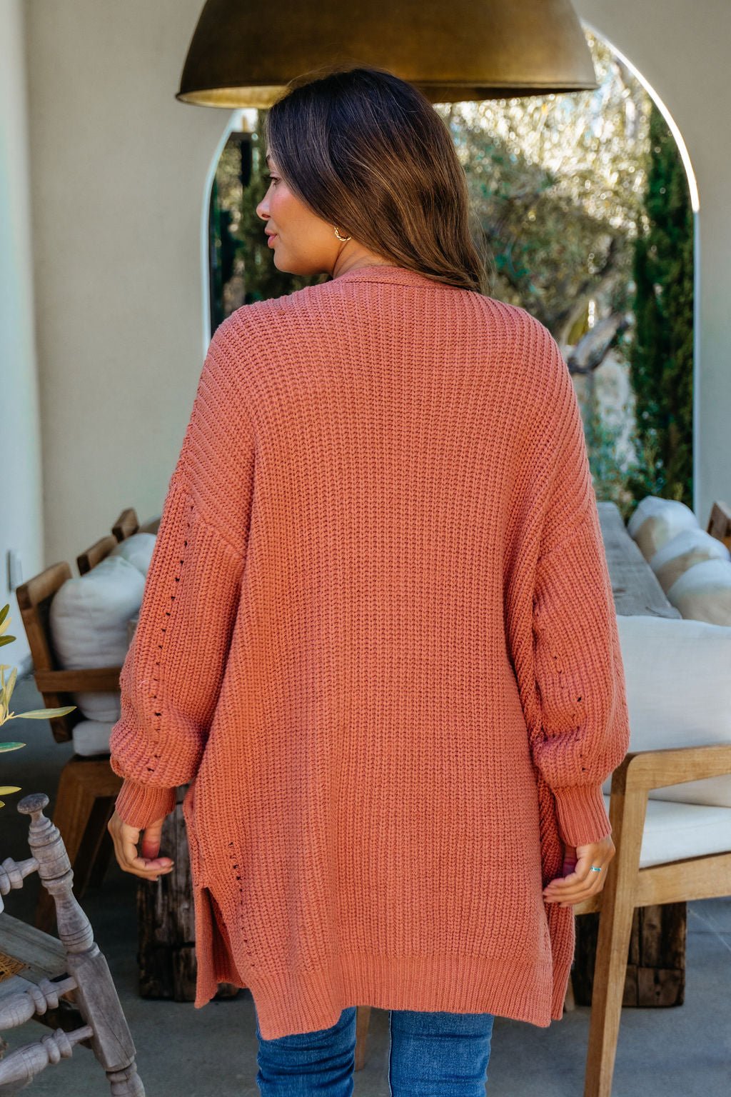 Woman with long brown hair wears a Terracotta Open Front Cardigan and blue jeans, standing indoors facing away.