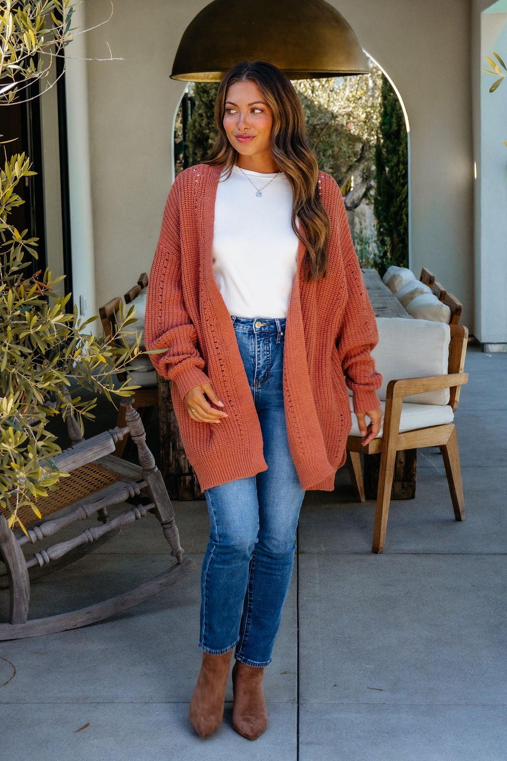 A woman wears a Terracotta Open Front Cardigan over a white top with blue jeans and brown boots, standing by a patio dining set.