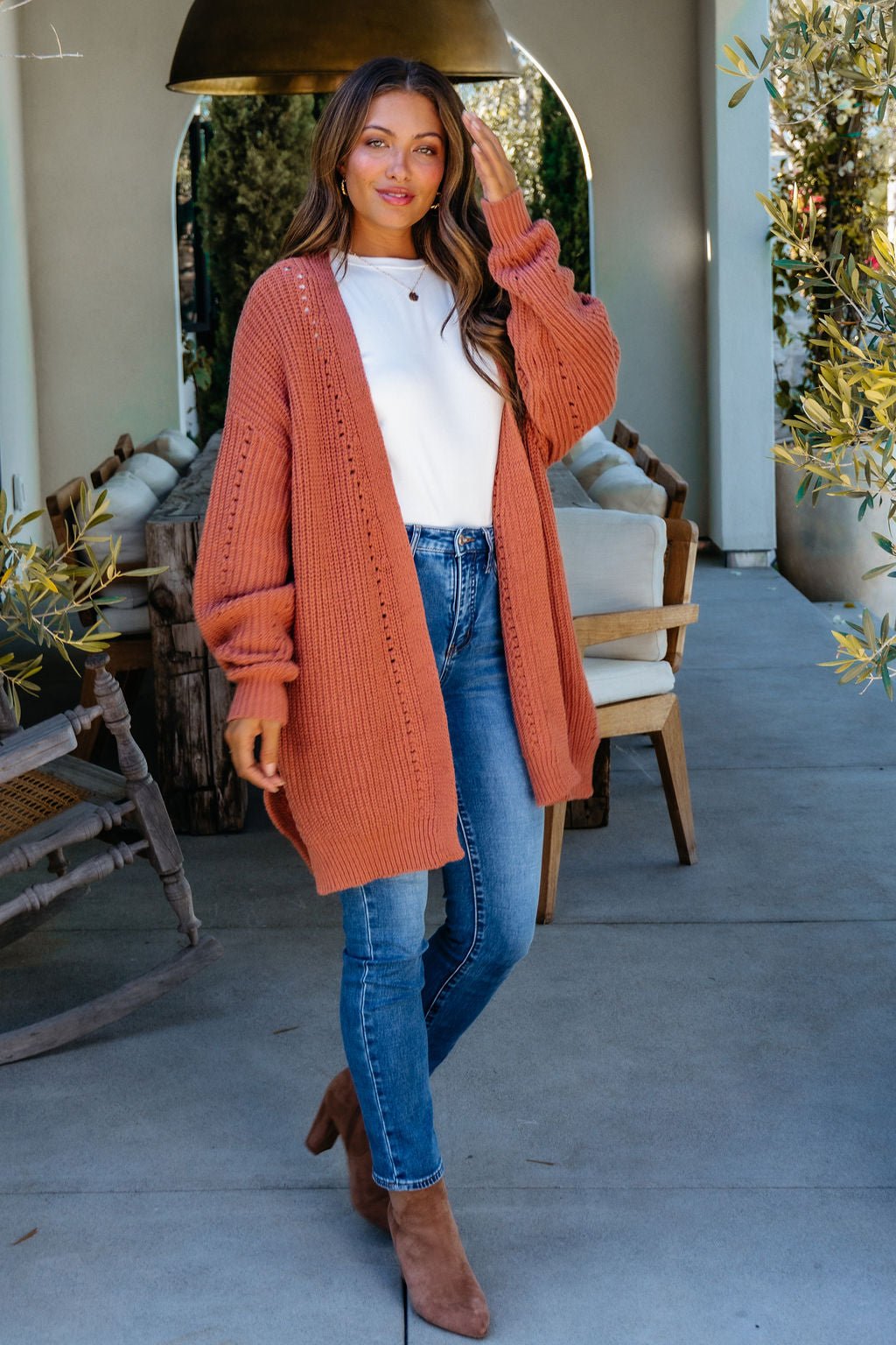 Woman outdoors wearing a Terracotta Open Front Cardigan over a white top with jeans and boots, smiling and touching her hair.