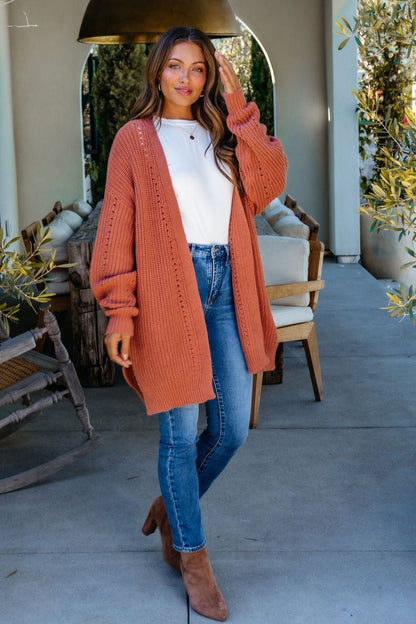Woman outdoors wearing a Terracotta Open Front Cardigan over a white top with jeans and boots, smiling and touching her hair.