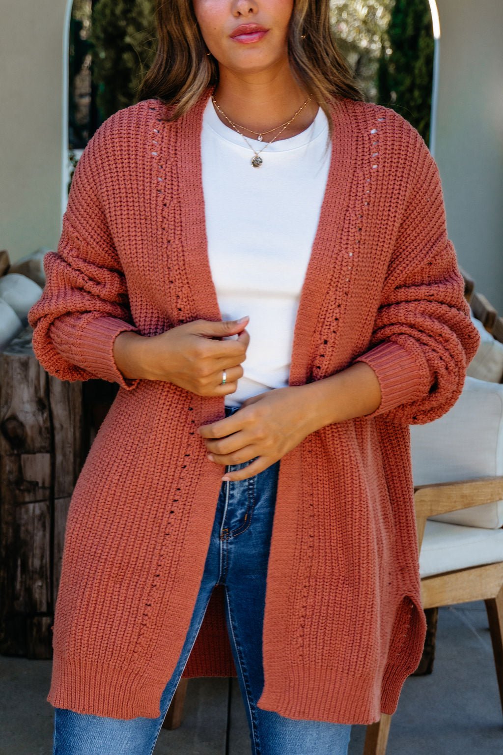 Woman wearing a Terracotta Open Front Cardigan over a white top and blue jeans, adjusting the cardigan indoors.
