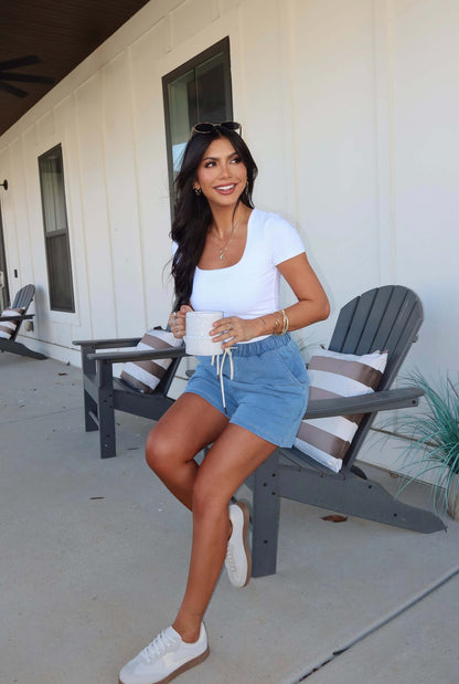 A woman smiles on a porch chair, holding a mug and wearing Terry Light Blue Lounge Shorts with a white top.