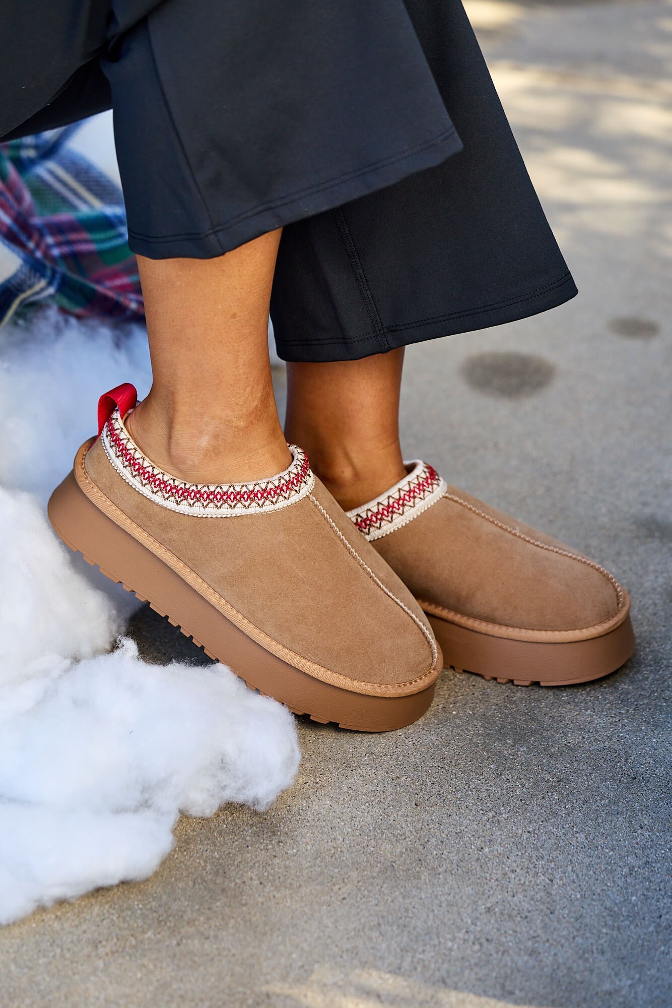 Person wearing Tessa Camel Platform Cozy Slippers with red and white trim, standing near a plaid blanket on pavement.