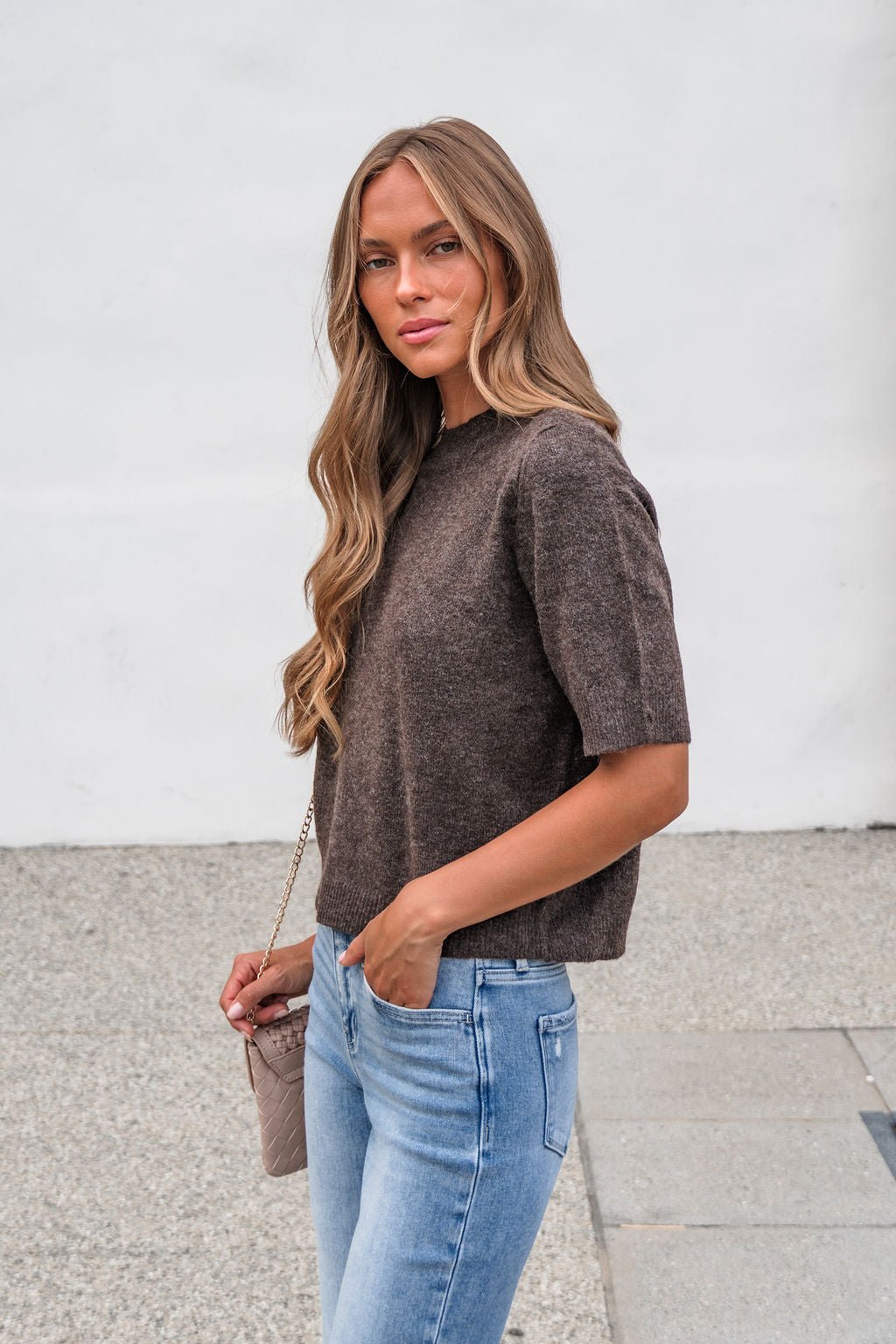 Woman with long wavy hair wears the Tessa Mock Neck Sweater in brown and blue jeans, holding a small purse outdoors.