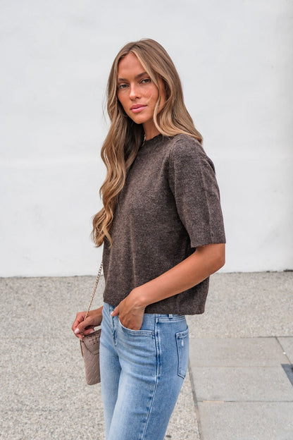 Woman with long wavy hair wears the Tessa Mock Neck Sweater in brown and blue jeans, holding a small purse outdoors.