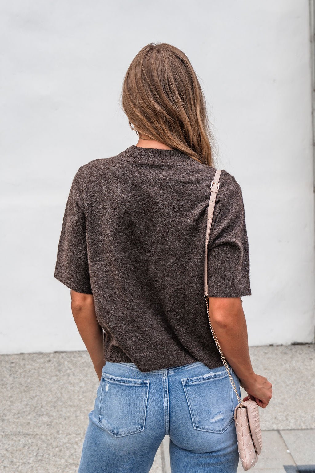 Woman with long brown hair in the Tessa Mock Neck Sweater - Brown and blue jeans, holding a beige purse, seen from behind.