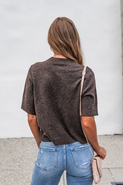 Woman with long brown hair in the Tessa Mock Neck Sweater - Brown and blue jeans, holding a beige purse, seen from behind.