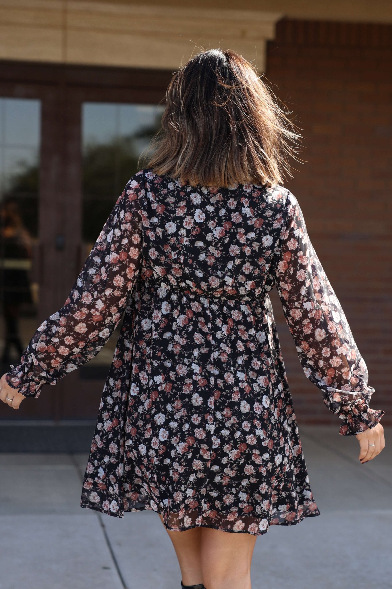 A woman in the Thalia Black Floral Print Mini Dress stands outdoors with her back to glass doors.