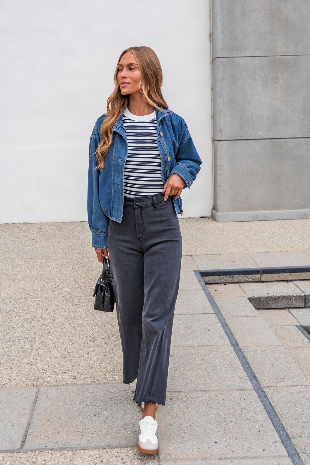 Woman in The Dakota Denim Jacket, striped shirt, dark pants, and white sneakers walks outside holding a small black bag.