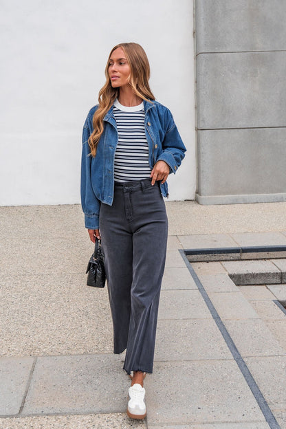Woman in The Dakota Denim Jacket, striped shirt, dark pants, and white sneakers walks outside holding a small black bag.