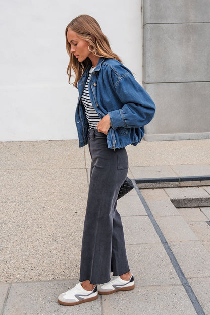 Woman wearing The Dakota Denim Jacket, striped shirt, and dark pants stands on pavement, looking down with a neutral backdrop.
