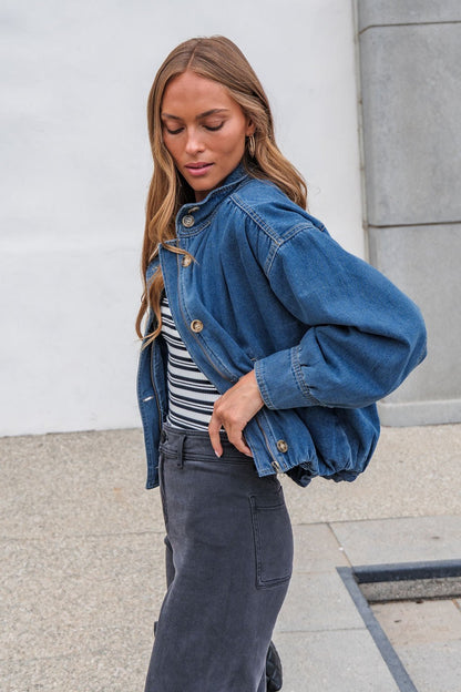 Woman in The Dakota Denim Jacket, striped shirt, and gray pants stands outdoors against a light background.