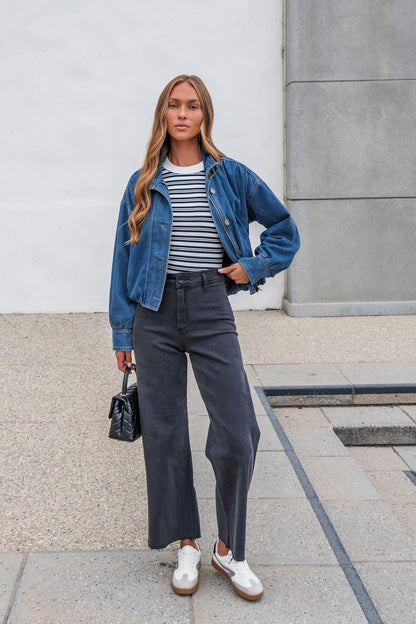 Woman in The Dakota Denim Jacket, striped shirt, wide-leg pants, and sneakers stands outdoors holding a small black handbag.