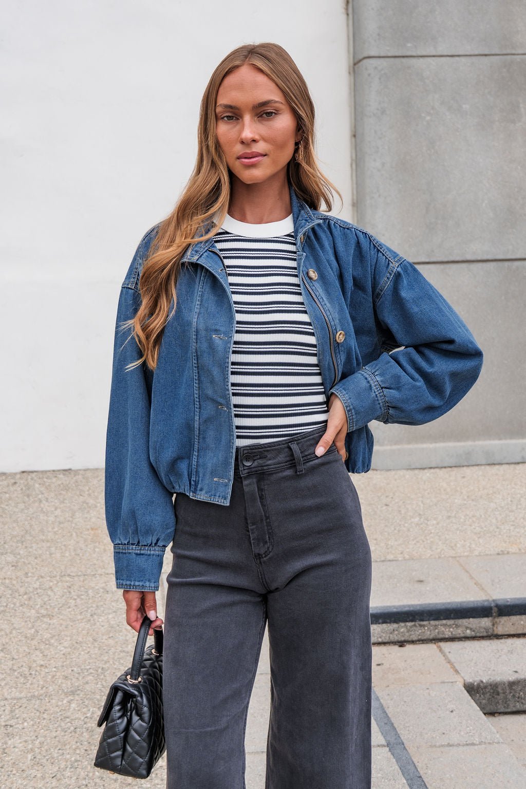 Woman in The Dakota Denim Jacket, striped shirt, and dark pants stands outside holding a small black handbag.