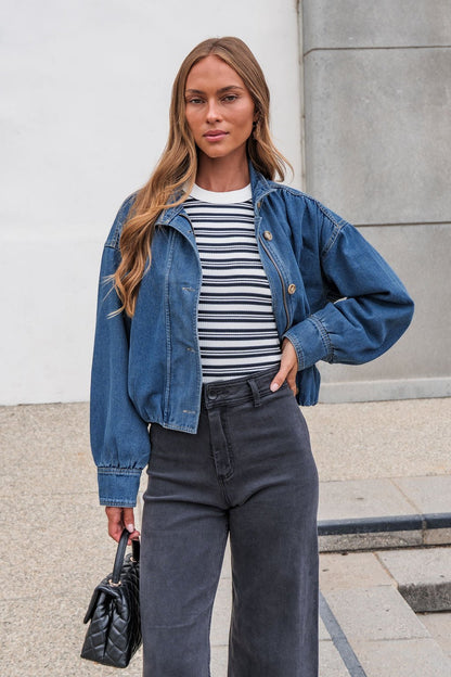 Woman in The Dakota Denim Jacket, striped shirt, and dark pants stands outside holding a small black handbag.