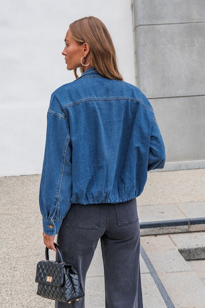 A woman with straight brown hair wears The Dakota Denim Jacket, dark jeans, and holds a black purse while standing outdoors.