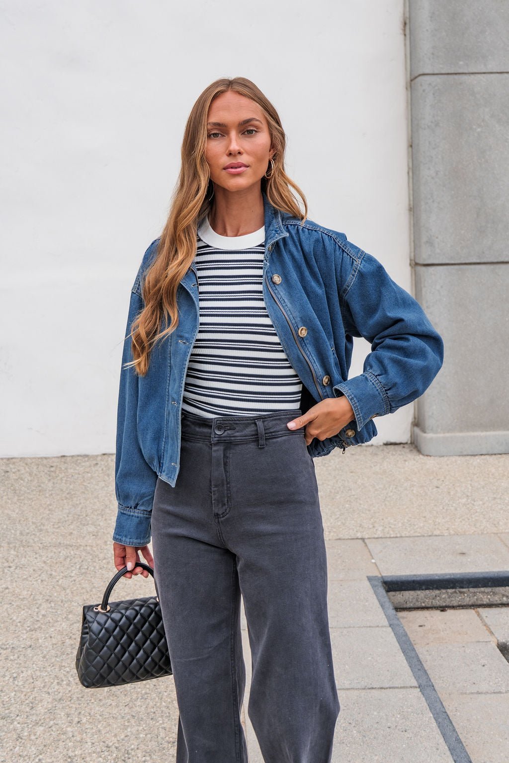 A woman in The Dakota Denim Jacket, striped shirt, and black jeans stands outdoors holding a small black quilted handbag.