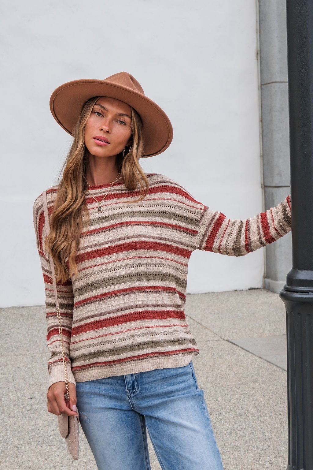 Woman wears The Delaney Striped Sweater - Rust, jeans, and tan hat outdoors, resting an arm on a black post and facing the camera.