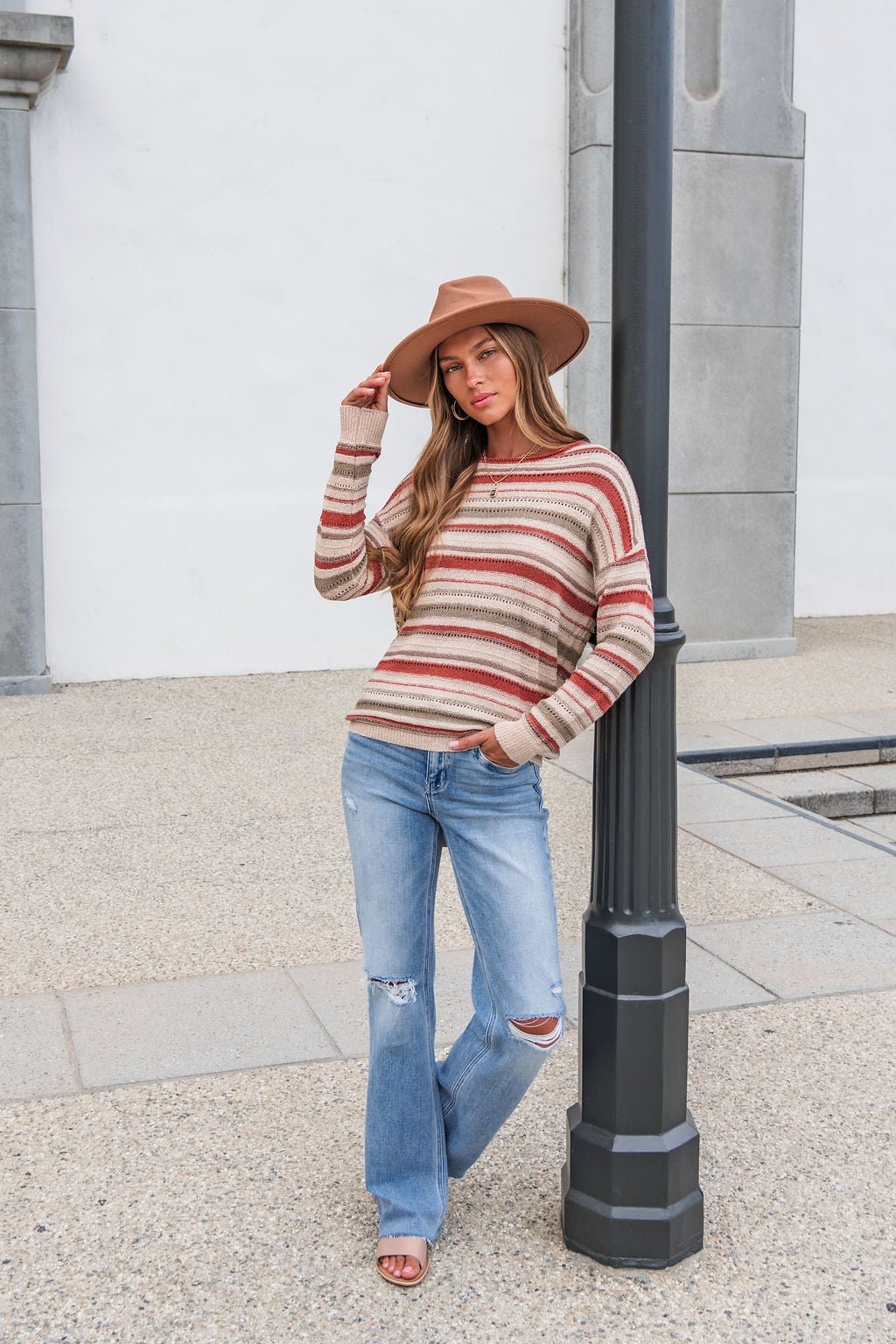 Woman in The Delaney Striped Sweater - Rust, ripped jeans, and hat leans on a lamp post on a concrete sidewalk outdoors.