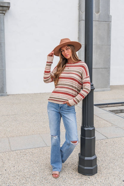 Woman in The Delaney Striped Sweater - Rust, ripped jeans, and hat leans on a lamp post on a concrete sidewalk outdoors.