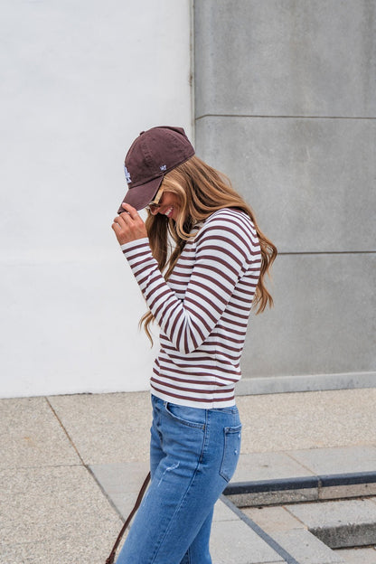 Woman in The Josephine Striped Top - Brown stands outside, smiling and looking down by a grey wall and pavement.