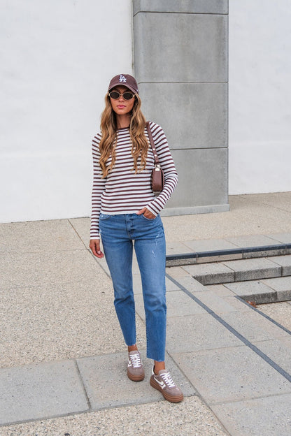 Woman in The Josephine Striped Top - Brown, blue jeans, brown sneakers, and hat stands on a concrete path holding a small bag.