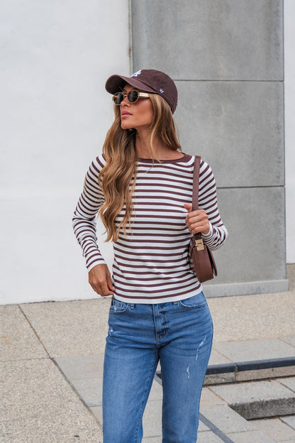 Woman in The Josephine Striped Top - Brown, blue jeans, sunglasses, and brown cap stands outdoors by a modern building wall.