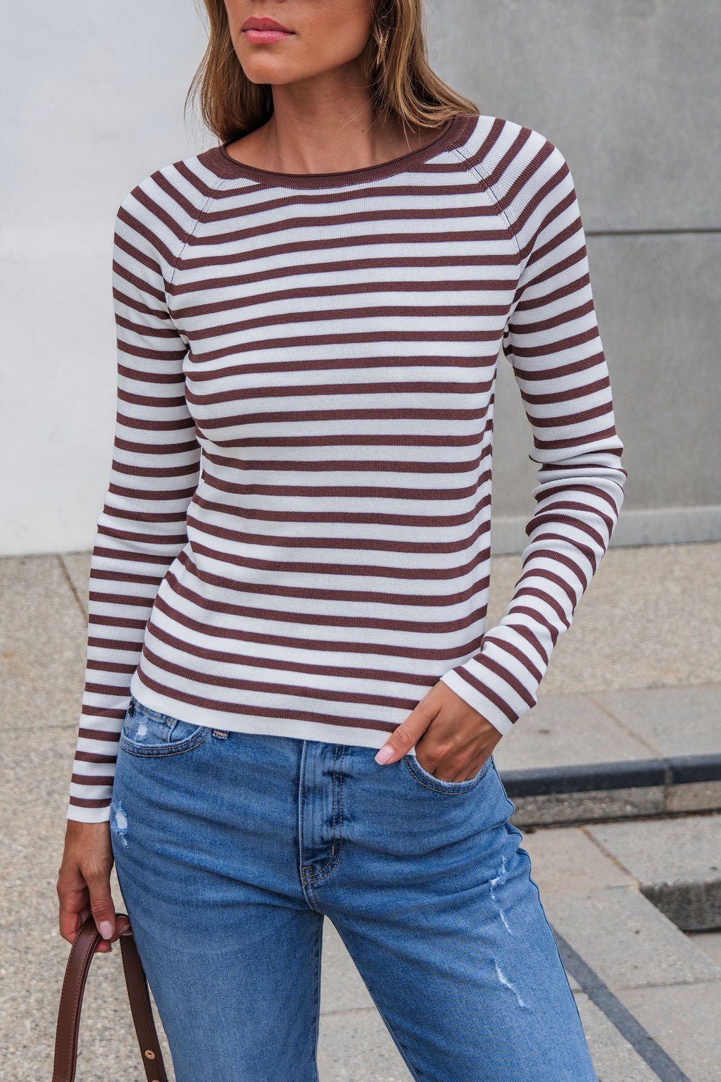 Woman wears The Josephine Striped Top - Brown with ribbed detail and blue jeans, standing outdoors with one hand in her pocket.