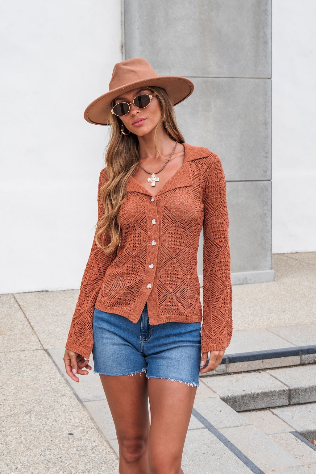 Woman in a brown hat, sunglasses, The Marisol Crochet Top - Terracotta, and denim shorts stands by a white wall and concrete steps.