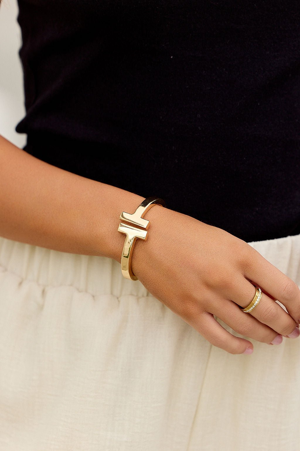 A hand wears The Maverick Cuff Bracelet - Gold and a gold ring, resting on a cream skirt with a black top.