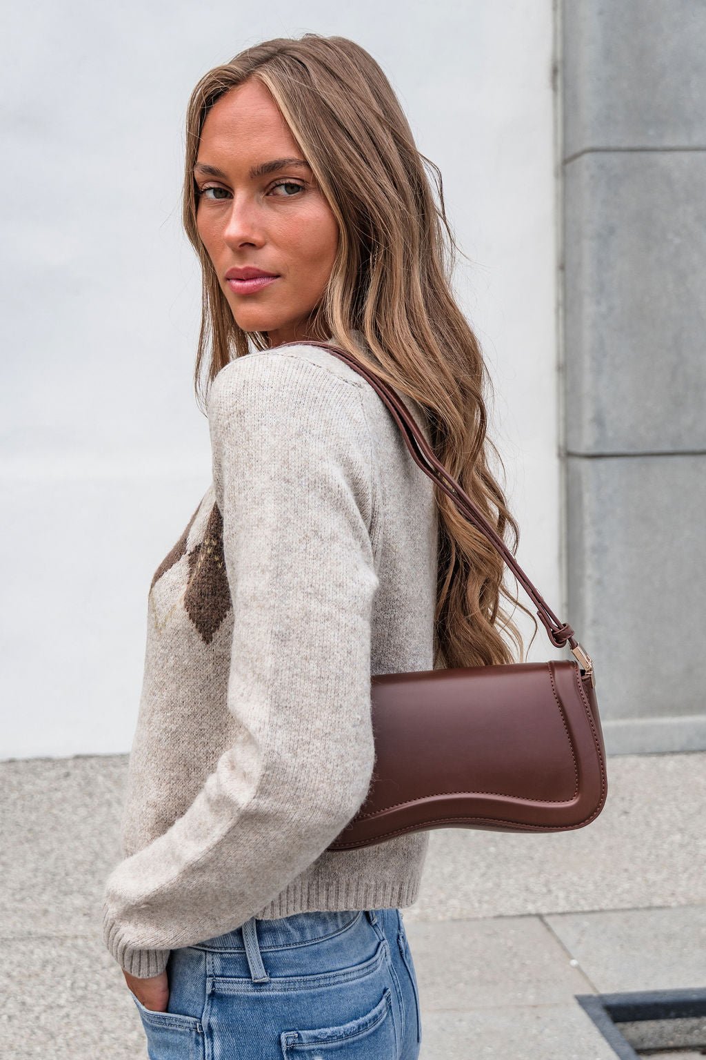 Woman with long hair in a beige sweater and jeans holds The Max Shoulder Bag - Brown by a building outdoors.