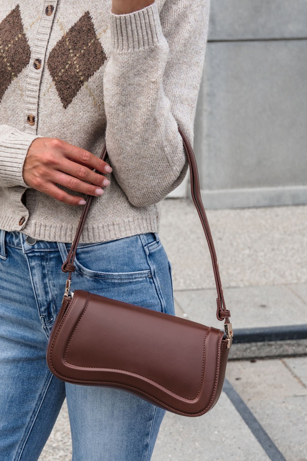 A person in jeans and a beige sweater carries The Max Shoulder Bag - Brown with an adjustable strap outdoors.