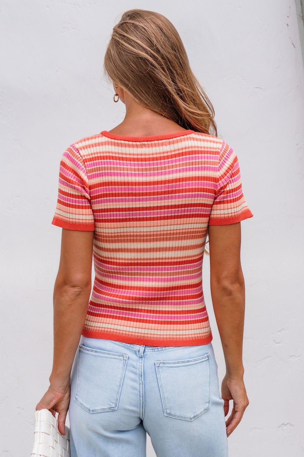 Woman facing away in The Piper Striped Tee - Pink, light blue jeans, and holding a white purse.