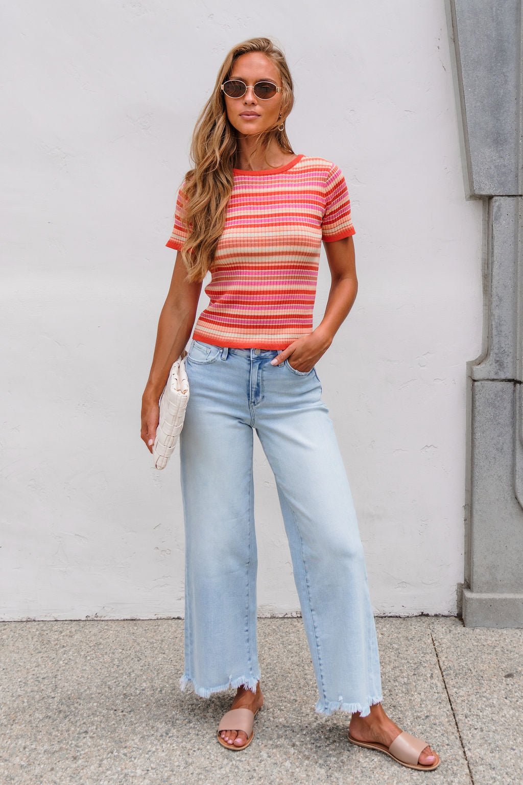 Woman in sunglasses wears The Piper Striped Tee - Pink, wide-leg jeans, sandals, and holds a white clutch by a white wall.