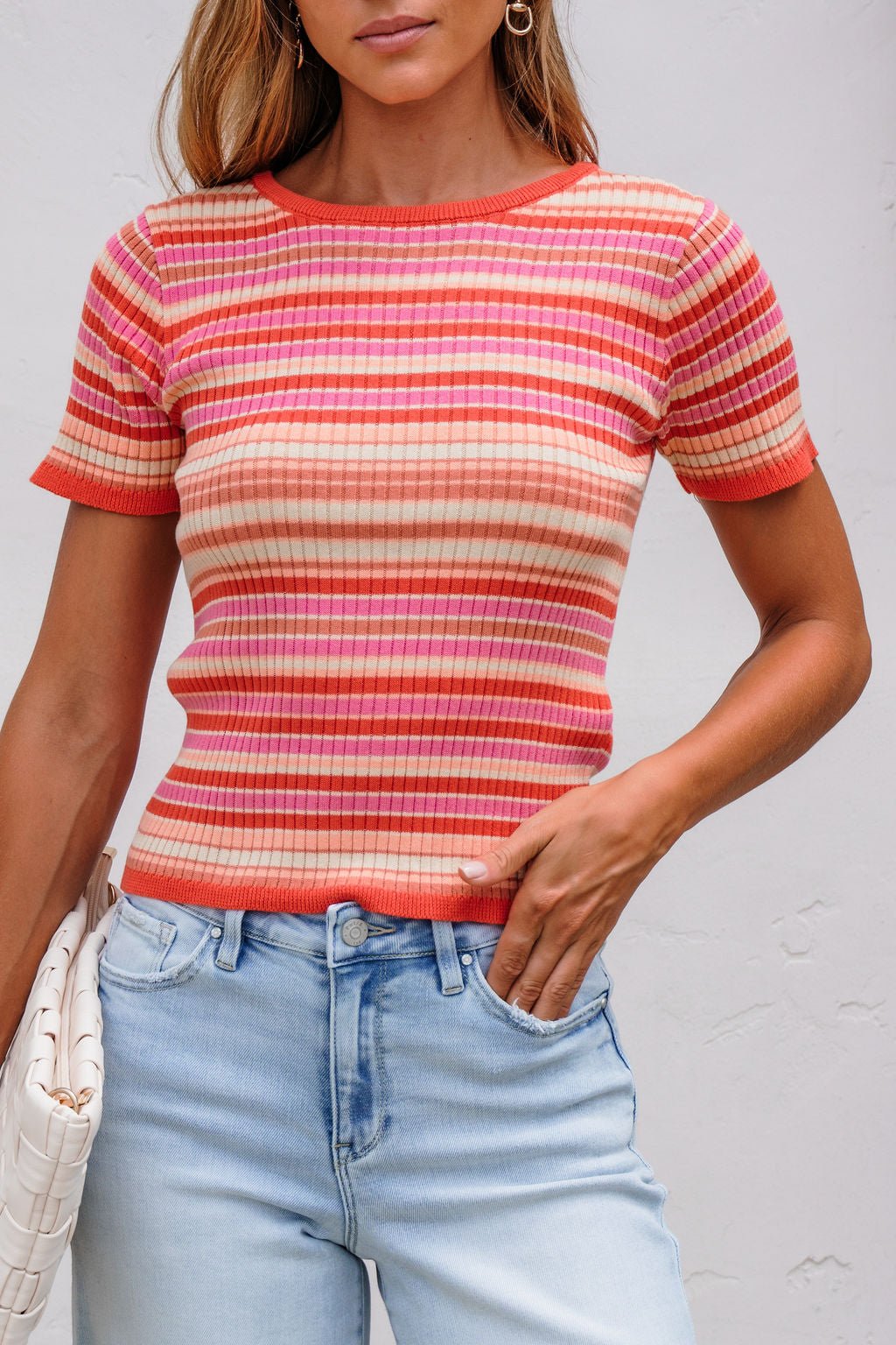 Woman in The Piper Striped Tee - Pink with light blue jeans, standing by a white wall.