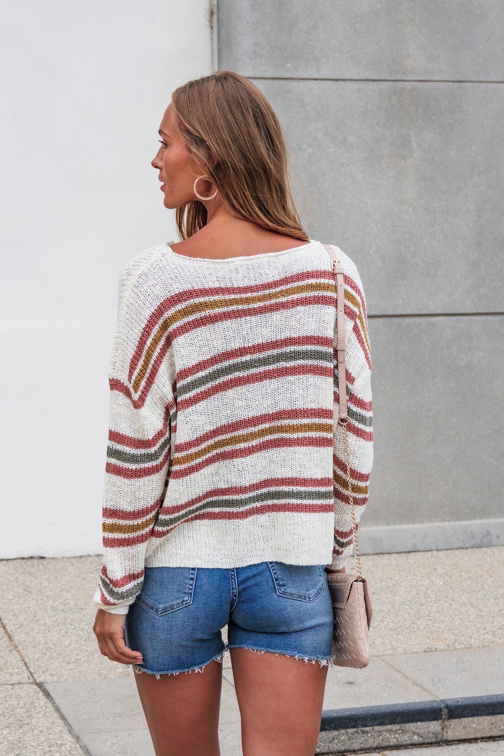 Woman with long brown hair wears The Reeves Striped Sweater - Red and denim shorts, standing outdoors facing away from the camera.