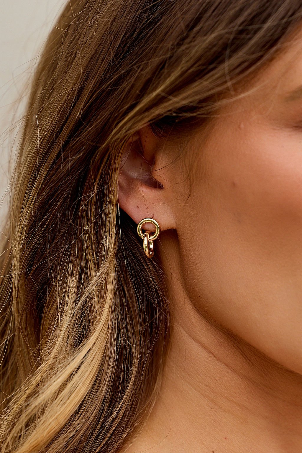 Close-up of a woman’s ear with brown hair, wearing The Roxie Double Hoop Earrings - Gold with a unique knotted double ring design.