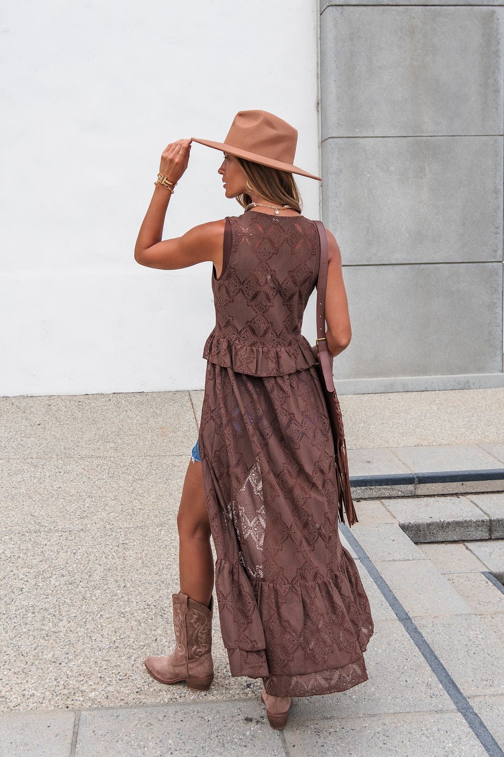 Woman in The Sorrel Lace Vest - Brown, hat, and cowboy boots stands outdoors facing away, holding her hat with one hand.