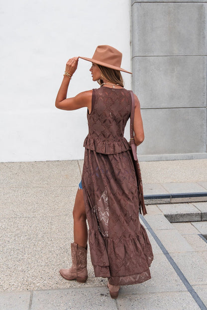 Woman in The Sorrel Lace Vest - Brown, hat, and cowboy boots stands outdoors facing away, holding her hat with one hand.