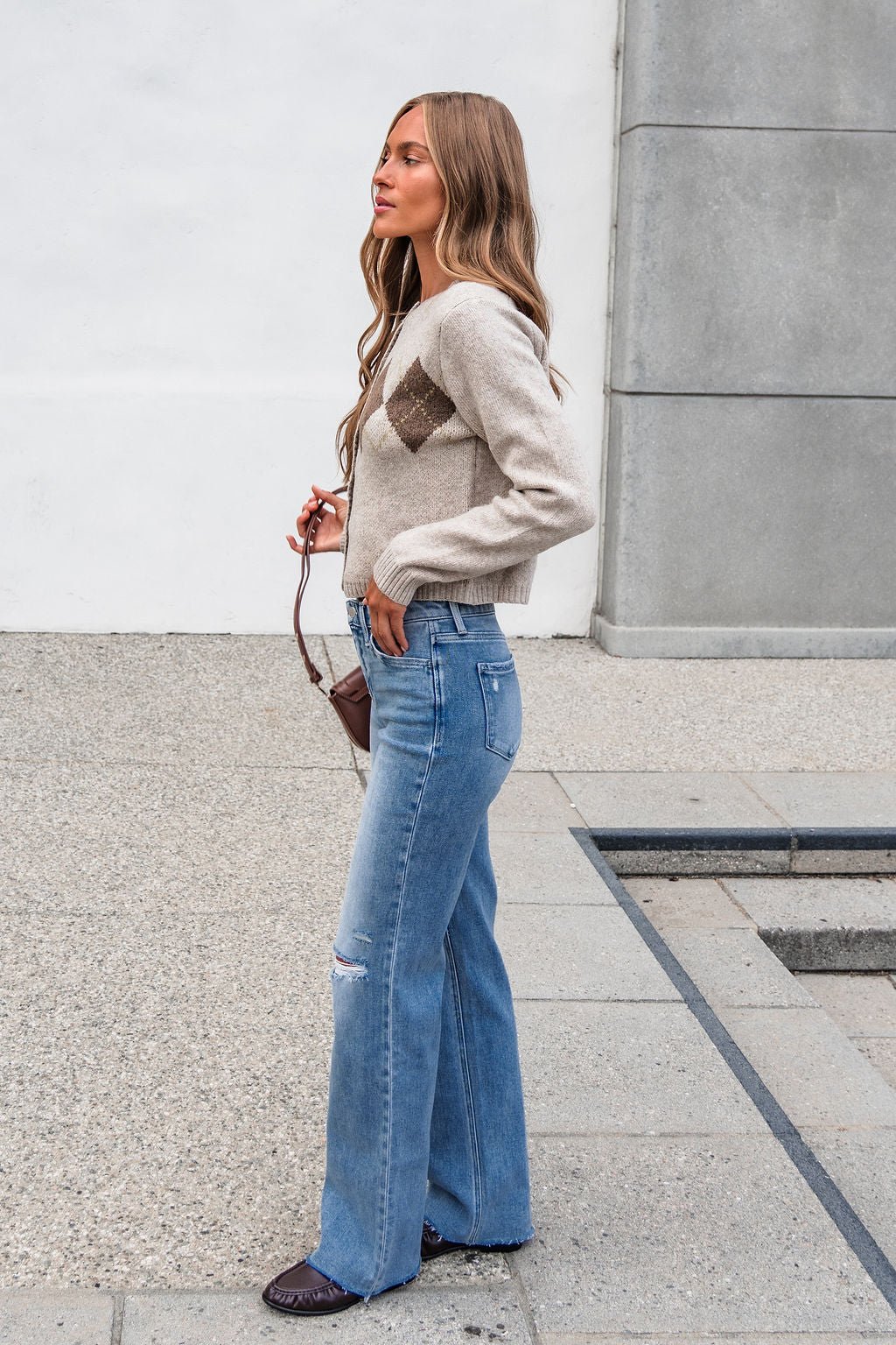 Woman in The Wynn Diamond Cardigan - Taupe and blue wide-leg jeans stands sideways outdoors, holding a small brown handbag.